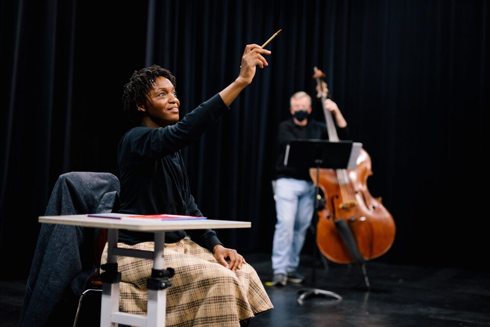 A+woman+sits+at+a+desk+pointing+up+with+a+pencil+while+a+man+plays+cello+behind+her Crédit photo : Diamond Edge Photography, Michelle Koebke
